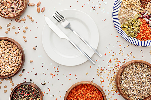 top view of empty plate with fork and knife near bowls with raw lentil, quinoa, oatmeal, beans, peppercorns and pumpkin seeds on marble surface with scattered grains