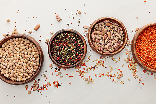 top view of wooden plates with chickpea, lentil, peppercorns, and beans near scattered grains on marble surface