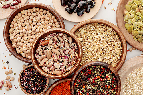 top view of wooden bowls with diverse beans, peppercorns, pumpkin seeds, oatmeal, quinoa and chickpea on white marble surface with scattered grains