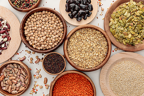 top view of wooden bowls with diverse beans, oatmeal, red lentil, peppercorns, pumpkin seeds and chickpea on white marble surface with scattered grains