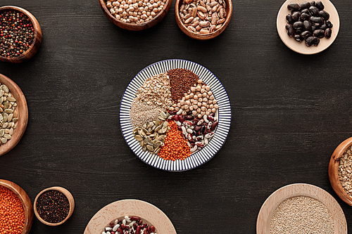 top view of striped plate with various raw legumes and cereals near bowls on dark wooden surface