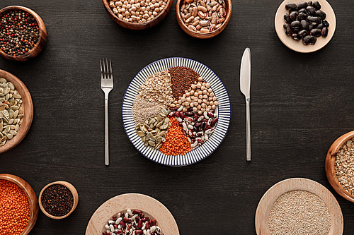 top view of knife and fork near striped plate with various raw legumes and cereals and bowls on dark wooden surface