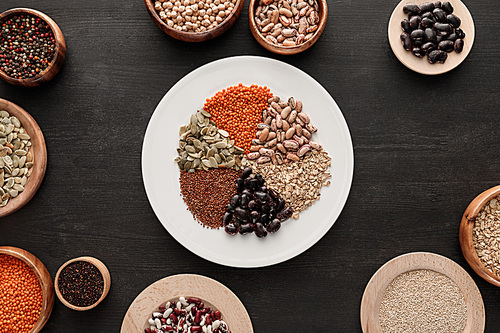 top view of white plate with various raw legumes and cereals near bowls on dark wooden surface