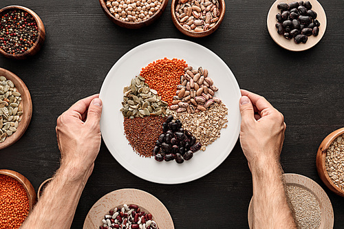 cropped view of man holding white plate with various raw legumes and cereals near bowls on dark wooden surface