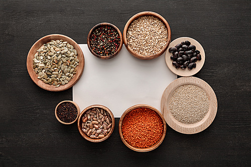 top view of blank white paper near various wooden plates and bowls with beans, cereals, spice and pumpkin seeds on dark wooden table
