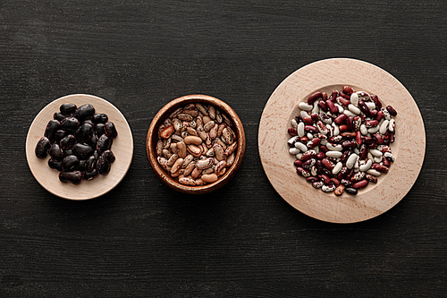 top view of bowl and plates with raw assorted beans on dark wooden surface with copy space