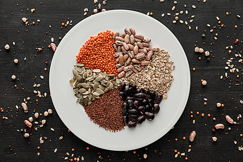 top view of white ceramic plate with raw assorted beans, cereals and seeds on dark wooden surface with scattered grains