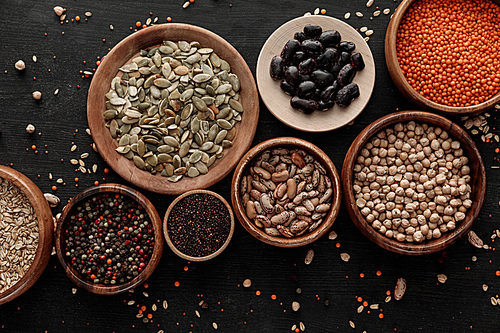 top view of wooden bowls and plates with raw assorted beans, cereals and seeds on dark surface with scattered grains
