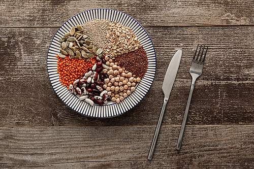 top view of fork and knife near striped ceramic plate with raw assorted beans, cereals and seeds on dark wooden surface with copy space