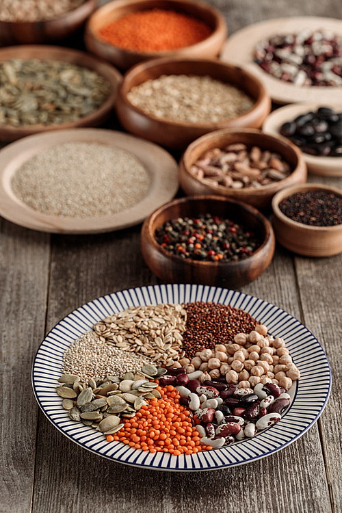 striped plate with lentil, beans, chickpea, cereals and pumpkin seeds near wooden bowls on table