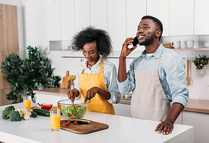 Woman mixing salad and boyfriend talking on smartphone in kitchen