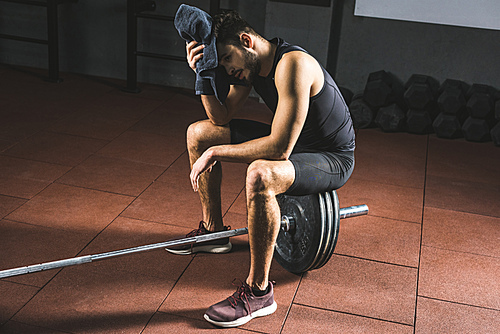 Tired sportsman wiping face by towel and sitting on barbell in gym