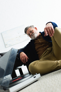 low angle view of smiling senior man in eyeglasses listening music with vinyl record player at home