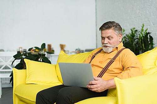 smiling bearded senior man using laptop while sitting on couch at home