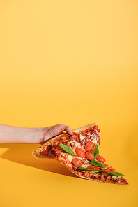 partial view of woman holding piece of pizza with cherry tomatoes in hand on yellow background