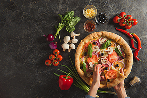 cropped shot of female hands and cooked italian pizza with fresh ingredients on dark tabletop