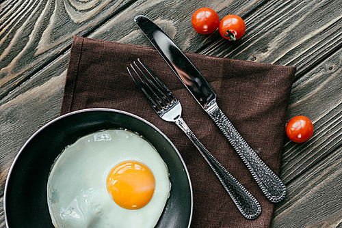Frying pan with egg and cutlery set on wooden table with tomatoes