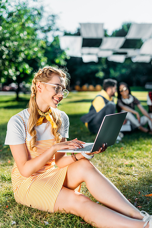 selective focus of young smiling woman using laptop and friends behind resting on green grass in park