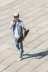 high angle view of handsome young skateboarder walking with longboard in hand and talking by phone