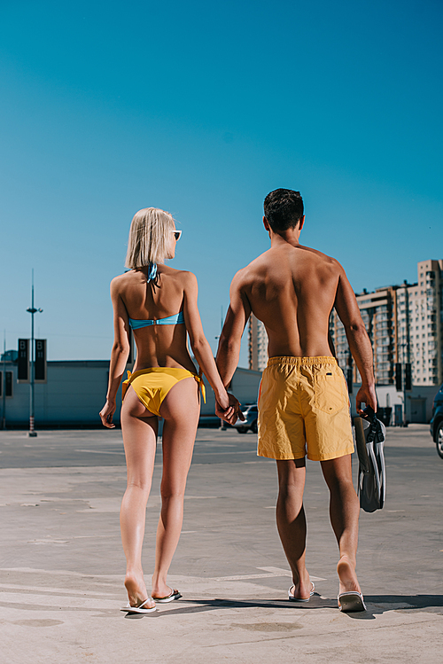rear view of young attractive couple in bikini and swimming shorts with flippers walking on parking