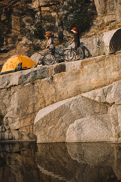 distant view of two male travelers  standing with mountain bikes near tent on rocky cliff over river