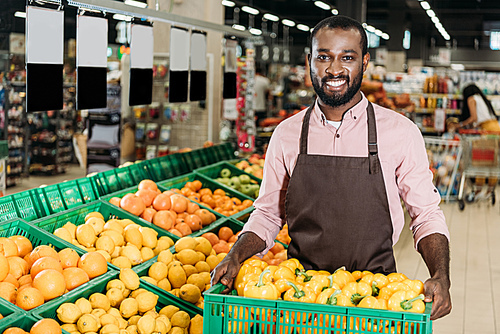 happy african american male shop assistant in apron holding box with bell peppers in grocery store