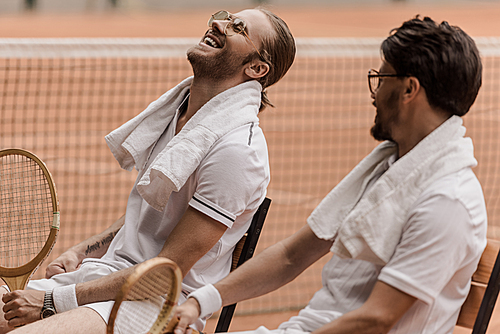 smiling retro styled tennis players sitting on chairs with towels and rackets at tennis court