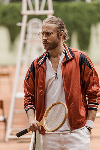 handsome retro styled tennis player standing with racket at tennis court and looking away