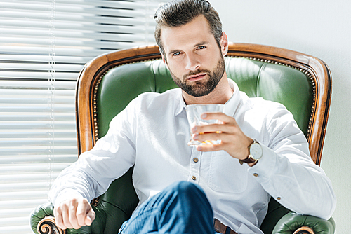 handsome elegant man holding glass of whiskey and sitting in green armchair