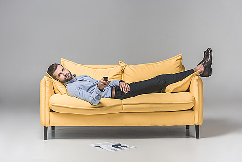 bearded man with remote control watching TV and resting on sofa with newspaper on floor, on grey