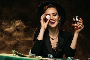 happy attractive girl in jacket and hat holding glass of whiskey at poker table and covering eye with casino chip