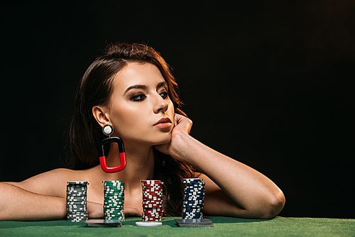 beautiful brown haired girl resting chin on hand and looking away, poker chips on table isolated on black