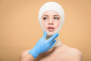 doctor in latex glove touching face of young woman with bandages on head and correcting lines isolated on beige