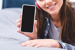 cropped shot of smiling young woman in headphones holding smartphone with blank screen while lying on bed