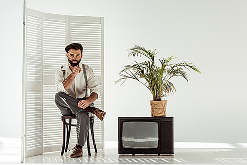 handsome bearded man sitting on chair near folding screen and tv in white room