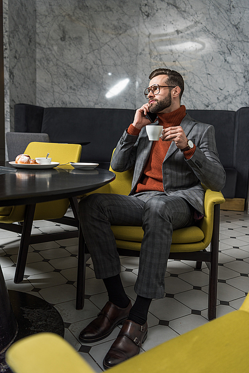 handsome man in formal wear talking on smartphone while drinking coffee in restaurant
