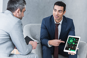 smiling young businessman pointing at digital tablet with business charts on screen and looking at male colleague during conversation