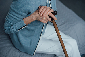 cropped view of senior woman sitting on bed with walking stick at home