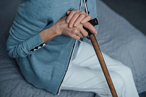 cropped view of senior woman sitting on bed with walking stick at home