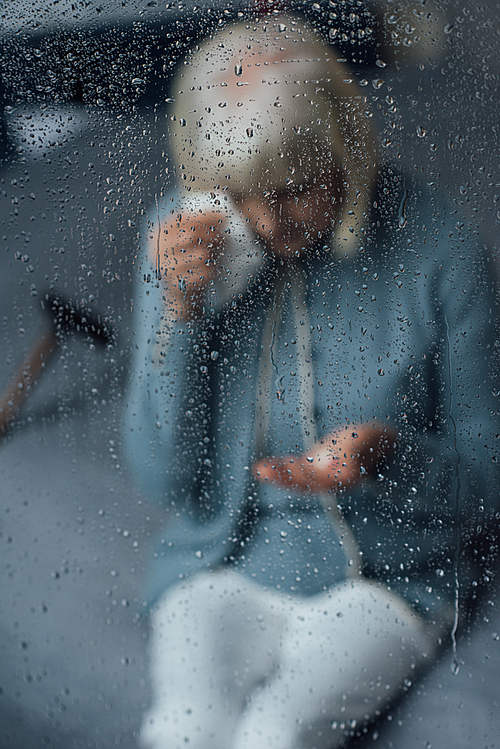 senior woman sitting on bed and wiping tears with handkerchief at home through window with raindrops