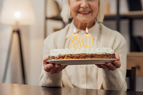 cropped view of smiling senior woman holding birthday cake with number 80 on top at home