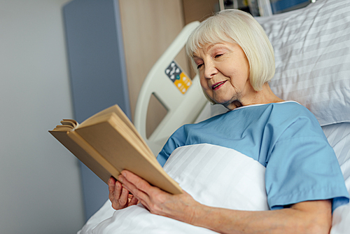 smiling senior woman with grey hair lying in bed and reading book in hospital