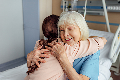 happy senior woman and daughter sitting on bed and hugging in hospital