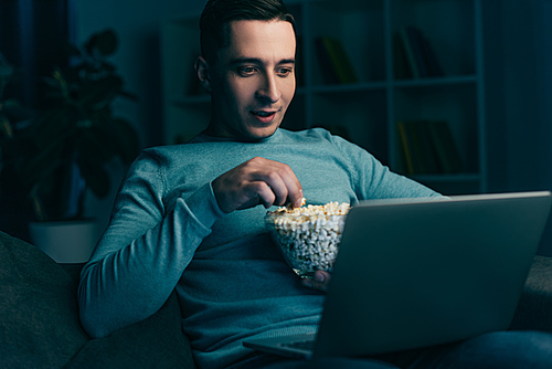 handsome man watching movie on laptop and holding bowl with popcorn at home