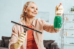 Stunning senior woman in rubber gloves cleaning window with glass wiper and spray