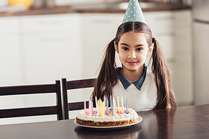 cheerful latin kid in party cap sitting near birthday cake with candles at home