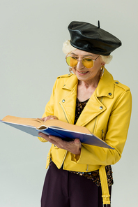 fashionable senior woman in yellow leather jacket reading book, isolated on grey
