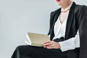 cropped view of senior lady with pearl necklace holding book, isolated on grey