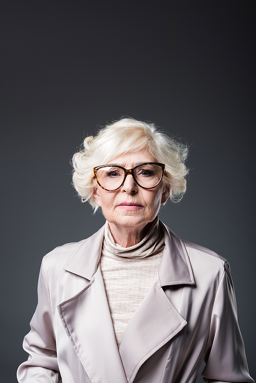 portrait of beautiful senior lady in trench coat and eyeglasses, isolated on grey