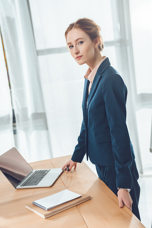 businesswoman in formal suit standing against table with laptop at office space